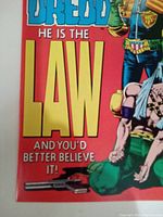 Close-up of Judge Dredd comic cover showing large yellow text and part of Judge Dredd standing with a captive on a red background.