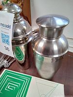 Two polished stainless steel cocktail shakers, one with strainer visible, placed on table with green cocktail mats underneath.