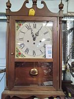 Front view of the wooden mantel clock showing clock face with Roman numerals, floral painted corners, glass door and woodwork details.