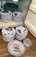 Two stacks of white porcelain plates with gold fruits, grapes and script design visible on top, each stack approx. 11 pieces