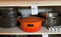 Shelf showing multiple large dark metal pots, the orange Le Creuset Dutch oven with missing lid pull, and a roasting rack.