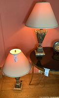 Two urn shaped lamps positioned against a coral wall, one lamp on the floor and one on a black round table, both with off-white lampshades with gold trim.