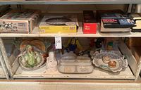 Shelf displaying ceramic turkey platter, white serving dish with rooster designs, ceramic figurine, paper mache clowns, and boxed items including taco rack and La Forme product
