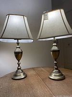 Pair of table lamps with brass bases and cream fabric trapezoid shades on a wood surface, showing overall look and condition.