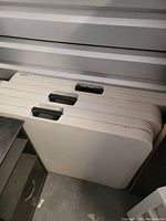 Three white folding tables stacked upright against a wall showing handles and edge sides.