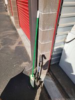 Photo of a green and white walking cane and black garden weeder tool leaning against a corner wall near a red roller door on asphalt.