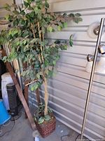 Artificial tree with braided trunk and green leaves situated against a wall with a woven basket base planter.
