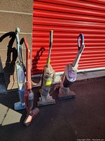 Four vacuums placed outdoors in front of a red garage door; one pink cordless stick vacuum, one orange upright vacuum, one Hoover G1 FloorMate SpinScrub upright vacuum, and one white/purple upright vacuum.