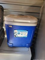 Blue rectangular Coleman ice chest cooler with white lid and integrated cup holders, showing some dirt and wear on the exterior.