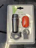 Front view of the bicycle light set in packaging showing black front headlight, orange rear taillight, and mounting parts.