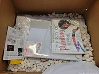 Box containing The Best of The Joy of Painting DVD, blank canvas, and printed art instruction materials, surrounded by packing peanuts.