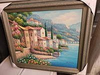 Full view of the framed oil painting showing the lakeside village landscape with houses, flowers, water and mountains.
