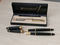 Four pens shown: Reflections boxed pen and three black pens with gold trim caps removed or placed aside, one fountain pen nib visible.
