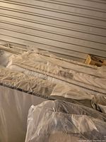 Photo showing a row of mattresses and box springs wrapped in protective plastic covers stacked against a wall.
