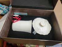 Photo showing white porcelain candle holder with attached red ribbon and scalloped porcelain candle cover with gold trim and embossed decorative knob inside original box.