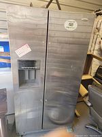 Front view of a stainless steel Whirlpool side-by-side refrigerator with water dispenser and stickers on the door. Shows signs of dirt and smudges.