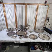 Full display of silver plated collection on white tablecloth showing variety of platters, pitchers, teapot, cups, and trays.