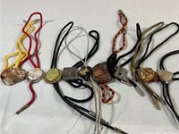 Collection of nine vintage bolo ties displayed in a row showing a variety of emblem shapes and cord colors.