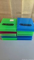 Stacked piles of Nintendo cartridge cases showing green, blue, and pink colored plastic cases with Nintendo logo on top, all empty and requiring cleaning.