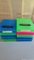 Stack of 10 empty Nintendo cases in blue, green, and pink, with Nintendo logo printed in black on the front. Some cases have white stickers with labeling.