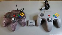 Two Nintendo 64 wired controllers and one memory card displayed on a flat surface. The purple translucent controller has a damaged analog stick and may be missing parts of the cord. The grey controller appears intact with a solid analog stick. The memory card is positioned between the controllers.
