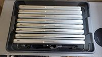 Top-down view of seven silver aluminum Apple MacBook Pro laptops and one MacBook laptop stacked horizontally in a plastic bin with additional parts beneath.