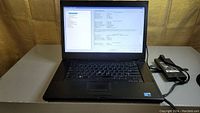 Dell Latitude E6510 laptop powered on and displaying BIOS system information screen, black laptop with power adapter beside it