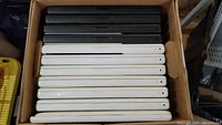 Top-down view of 11 MacBooks stacked side by side in a box, showing 6 white and 5 black units with glossy plastic bodies.