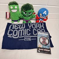 Front view of all items: three Marvel plush toys (two Hulk variations and one Captain America), navy blue New York Comic Con 2016 T-shirt with white text, Black Widow Avengers member exclusive patch in plastic sleeve.