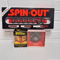 Photo showing Spin-Out brain teaser puzzle with black plastic frame and red sliding knobs in original box plus Inversion Puzzle and Curious Cross puzzle boxes below it.