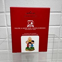 Front view of ornament box with red background and image of Peanuts characters Charlie Brown, Linus, Lucy, and Snoopy on grassy base. Text highlights '70 Years' and 'You're a Good Man, Charlie Brown!'