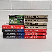 Photo showing variety of sealed cassette tapes including Maxell XLII 90, Maxell UR 90, Memorex dB C90 and C60, Sony HF 60, and Fujifilm DVC tapes