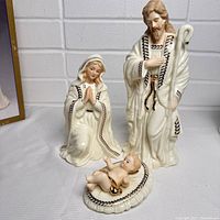 Three jade porcelain nativity figures of Mary, Joseph, and Baby Jesus arranged on table against a white brick wall.