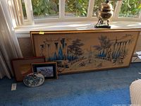 Wide view of four framed artworks placed on carpet near window with sunlight, showing diversity of art including large piece and smaller framed pieces