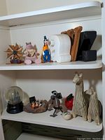 Two shelves with various decorative items including Goofy figurine, handcrafted wooden llamas, white ceramic giraffe bookends, wooden windmill, snow globe, and small wooden and metal sculptures.