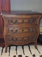 Front view of wood chest of drawers showing three curved drawers with ornate metal handles and keyhole escutcheons, and visible scratches on surface.