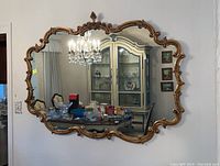 Full view of large ornate wooden framed wall mirror mounted on a white wall, reflecting crystal chandelier and dining set.
