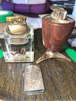 Overall view of three lighters: sterling silver engraved lighter, Ronson lighter with wood and metal tripod base, and plexiglass lighter with miniature figures inside.