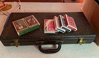 View of vintage mahjong case with textured dark brown finish, staple handle, and multiple decks of playing cards including some loose cards and a box of music playing cards placed on top.