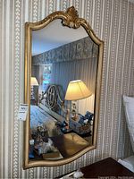 Front view of the gold framed wall mirror hanging on striped wallpaper near bedside table and lamp.
