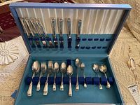 Photo of the open flatware storage box showing various silverplate forks, spoons, and knives arranged neatly in blue velvet slots.
