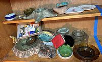 Overview of multiple ashtrays of glass, ceramic, and metal on wooden shelf