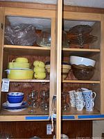 Wooden shelf cabinet with vintage yellow ware dishes, glass cake stands, brown glass bowls, stemware glasses, and ceramic mugs and bowls.