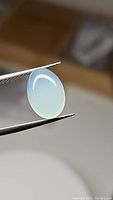 Oval cabochon cut Arctic Blue Chalcedony gemstone held by tweezers, showing its translucency and pale blue color.