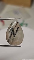 Close-up image of pear-shaped black rutilated quartz cabochon held between tweezers showing its clarity and black needle inclusions.