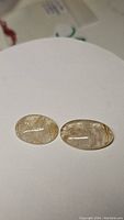 Two oval cabochon cut golden rutilated quartz stones displaying natural golden needle inclusions, laid flat on white background.