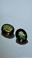 Two oval cut green sapphires placed on a white surface showing their color and cut from different angles.