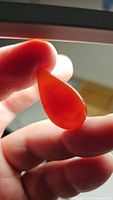 Photo showing a close-up of a pear-shaped deep orange carnelian cabochon held between fingers with light shining through, highlighting its translucency and natural color.