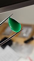 Close-up photo of a pear-shaped, deep green onyx cabochon gemstone held in tweezers against a blurred background.