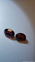 Pair of oval cut dark reddish-purple garnet gemstones placed flat on white background showing surface and some internal clarity features.
